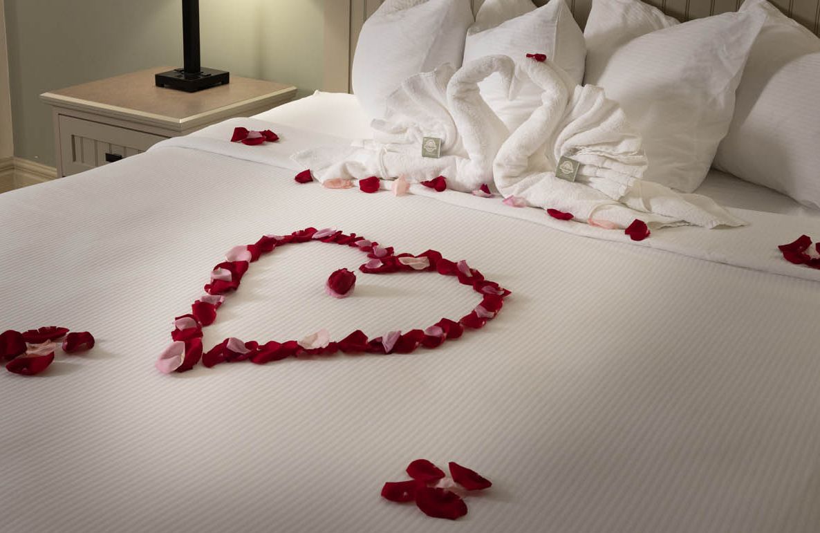 A perfectly manicured bed with rose petals int he shape of a heart, with two towels made to look like kissing swans
